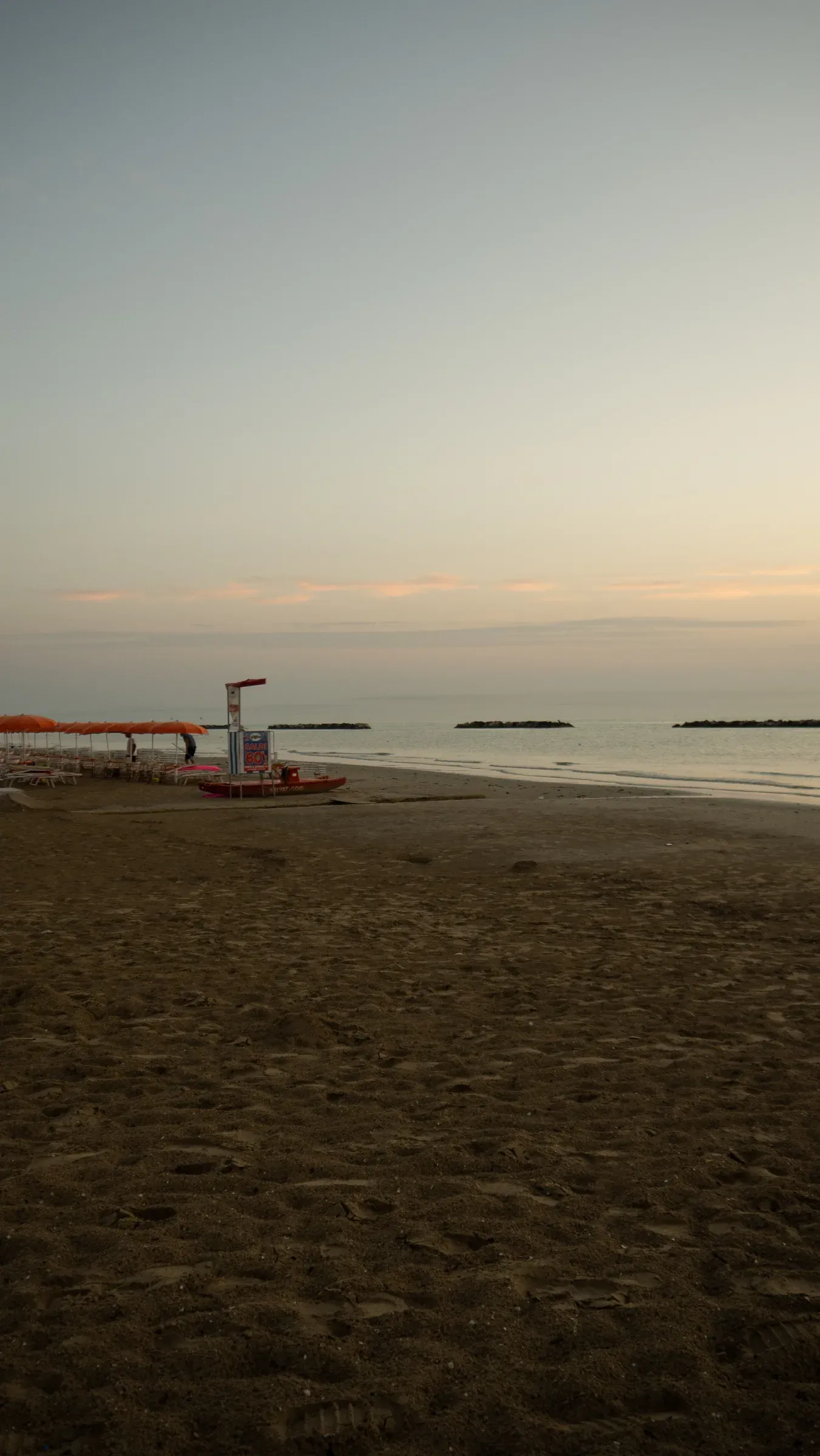Empty Adriatic beach at dusk with closed orange umbrellas, a lifeguard tower, and calm water