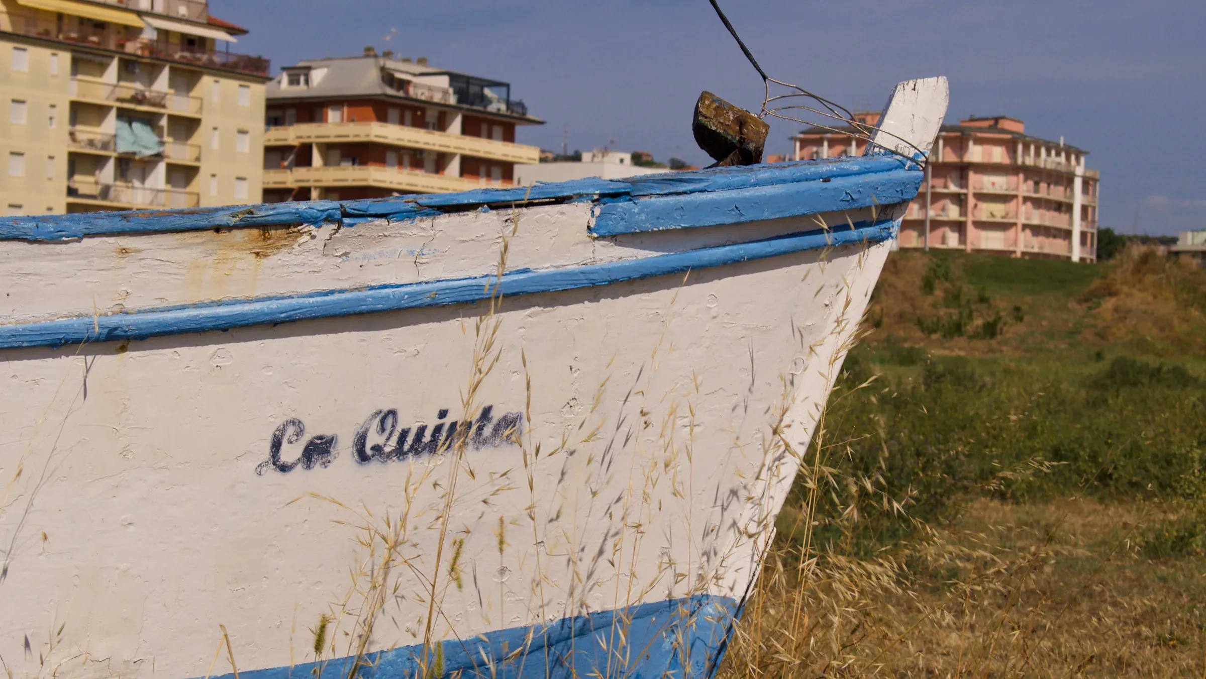 Weathered fishing boat named La Quinta resting on grass with coastal apartment buildings behind