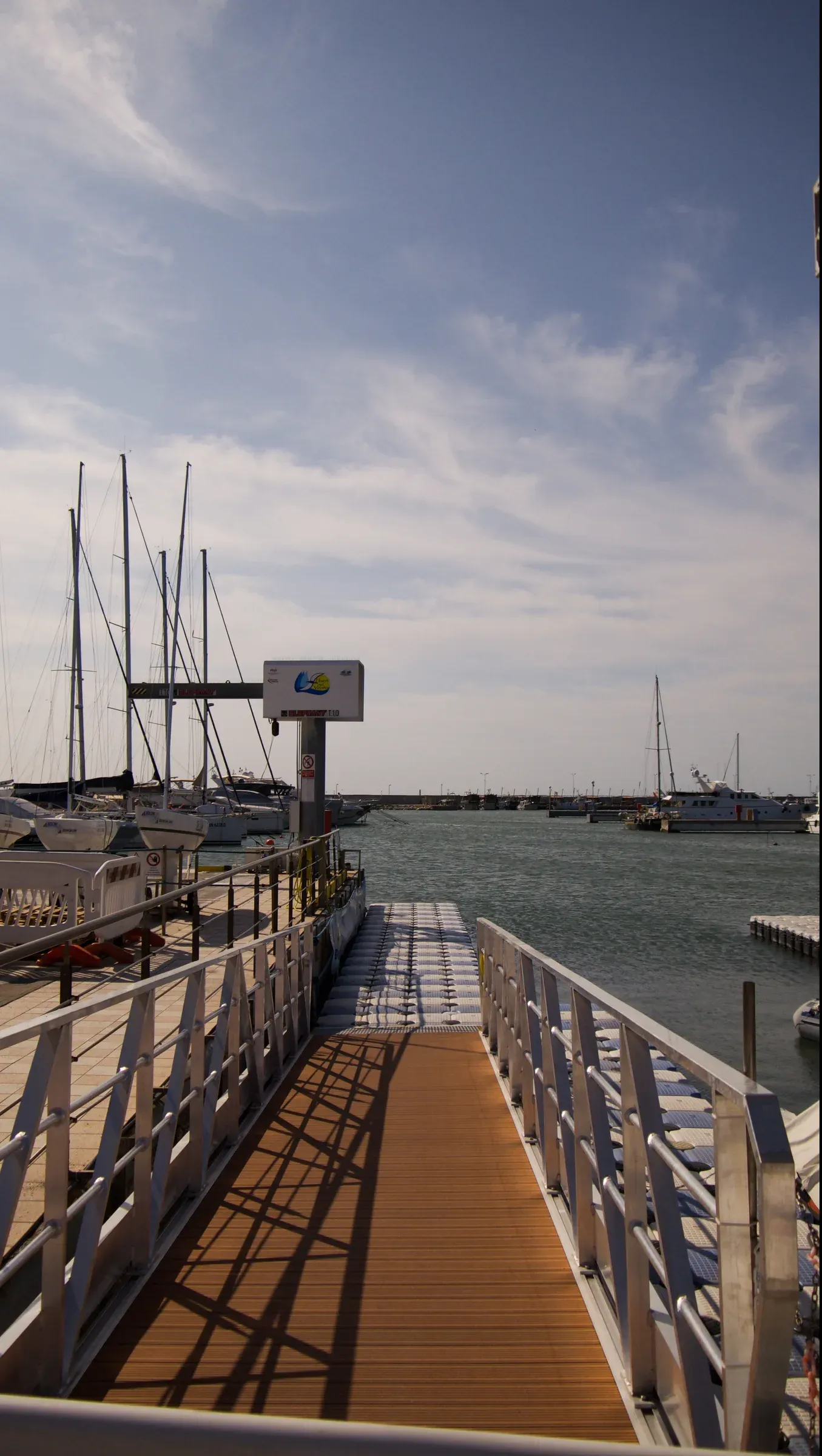 Long shadows on a wooden gangway leading toward sailboat masts in the harbor