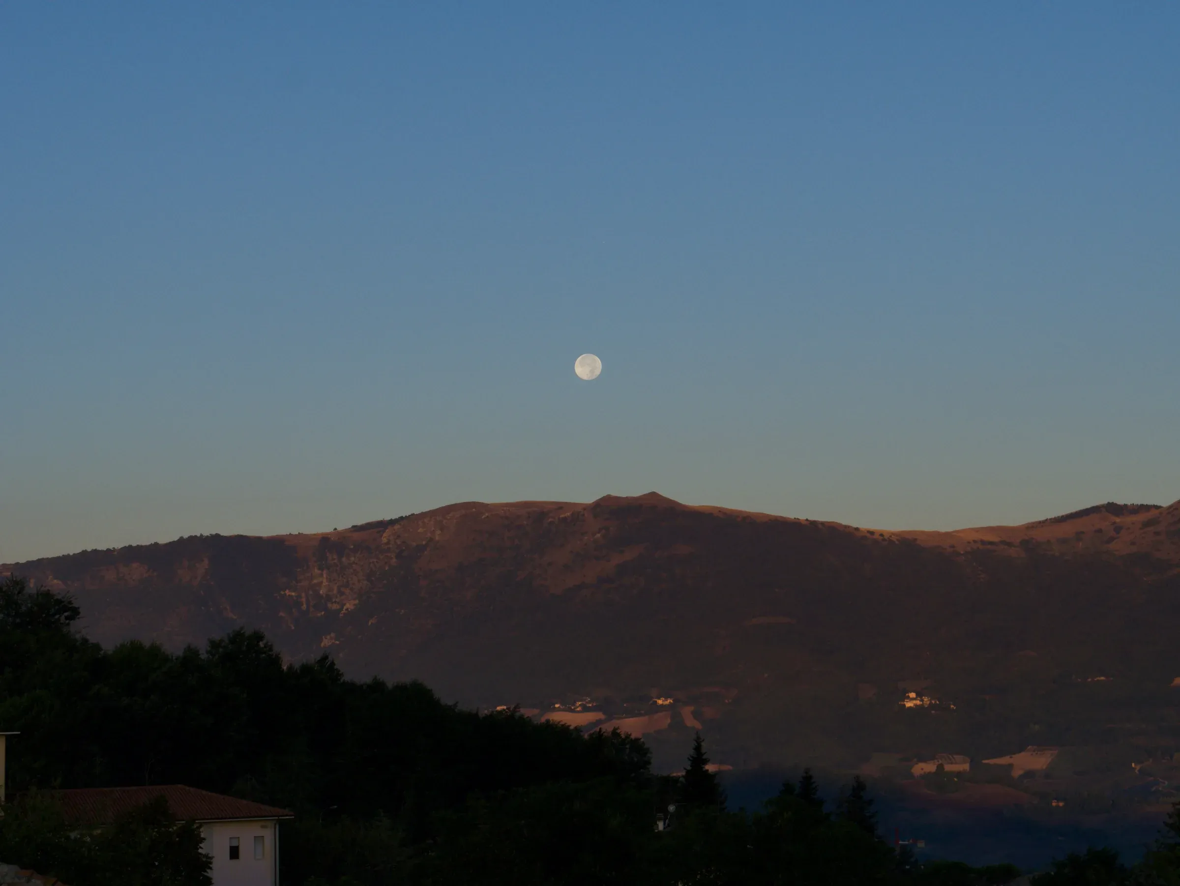 Full moon in a blue dawn sky above sunlit Marche hills and rooftops